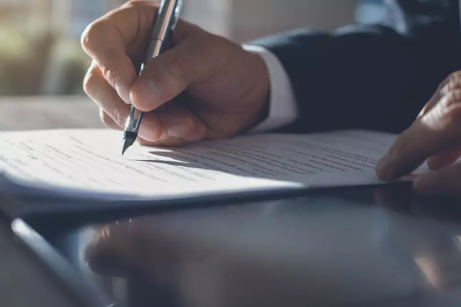 Dłoń w garniturze podpisująca dokument piórem wiecznym, druga dłoń wskazuje miejsce podpisu